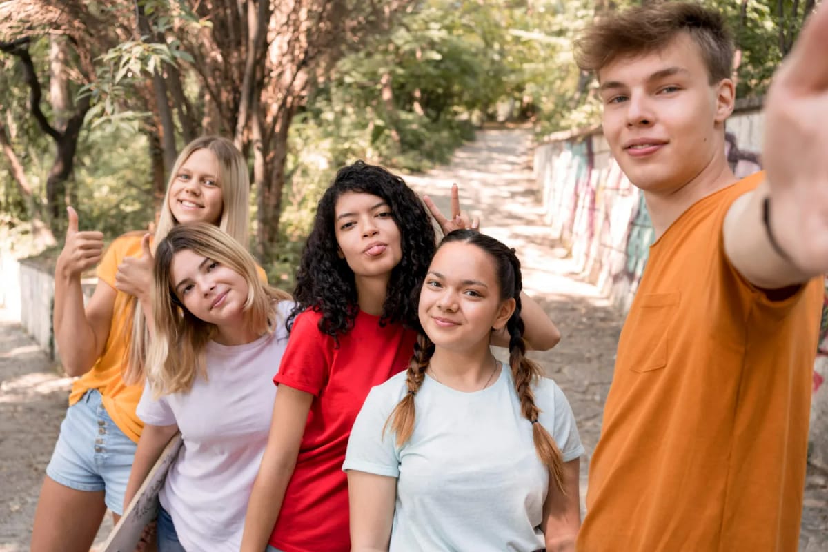 Group of five happy teenagers posing outdoors, one making a peace sign, another giving thumbs up.